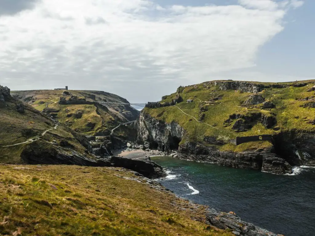 Standing on a grass hill looking down to the sea on the walk out of Tintagel. The sea is surrounded by grass topped hills and cliffs. There is a small beach enclosed by the cliffs.