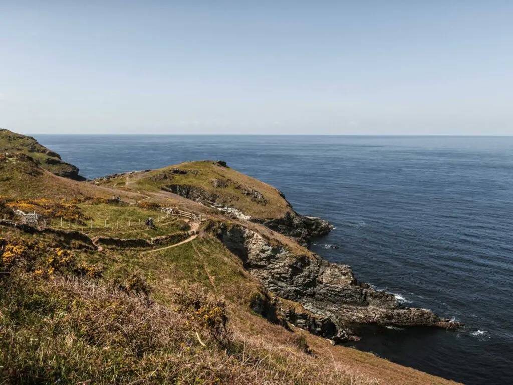 A grassy undulating landscape, with a trail running through it. The land forms cliffs as it meets the blue sea.