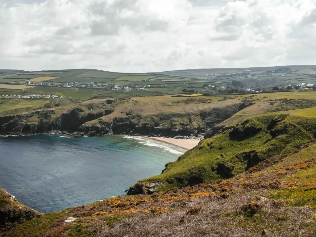 Looking across the grass covered hill towards the beach cove of Bossiney. There is a cliff on the other side of the beach leading to an undulating landscape.