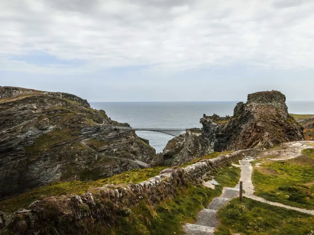 Steps leading down alongside a brick wall on the walk towards Tintagel island. There is a high bridge from the top of the island onto the hill on the mainland. The sea is behind.