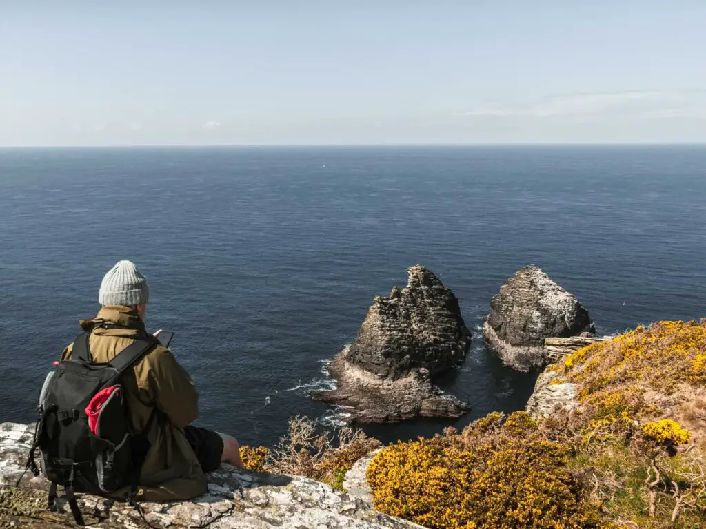 A man sitting on a rock looking down to two rocks in the blue sea on the coastal walk from Tintagel to Bossiney.