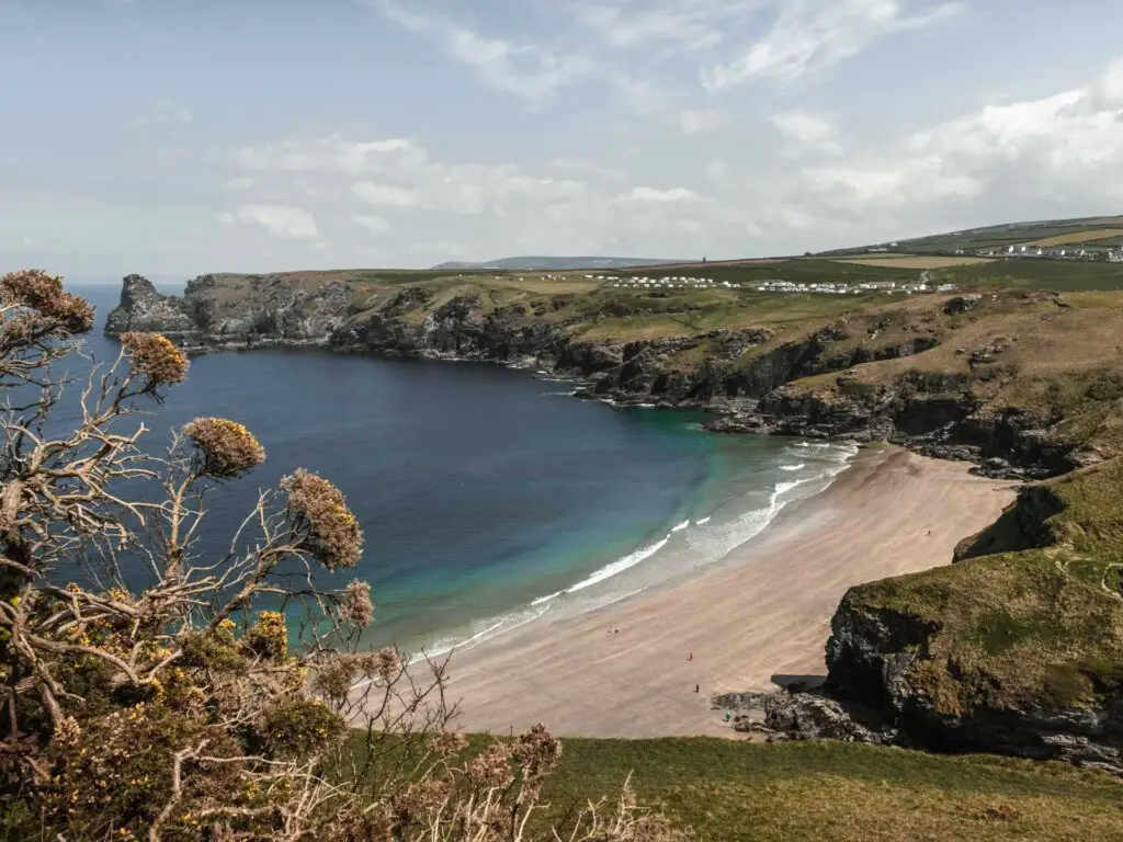 The pale sandy beach of Bossiney Cove on the walk from Tintagel. The blue sea becomes turquoise at it meets the beach. It is surrounded by craggy land and cliffs.