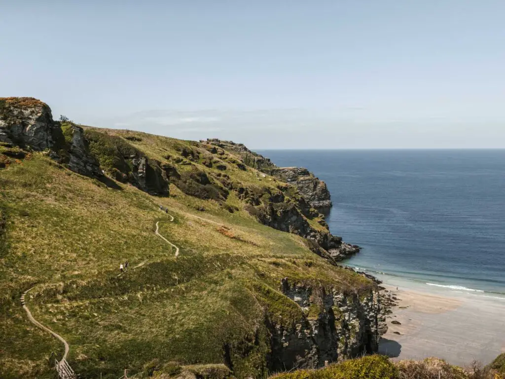 A trail running across the side of a grass topped hill on the walk to Bossiney from Tintagel. The hill leads to a sheer cliff drop onto a sand beach. The sea is blue.