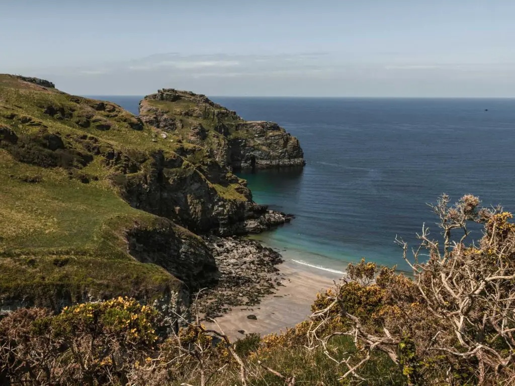 The sandy beach of Bossiney Bay enclosed by craggy cliffs on the walk from Tintagel. The top of the cliff is rugged and hilly and covered in grass.