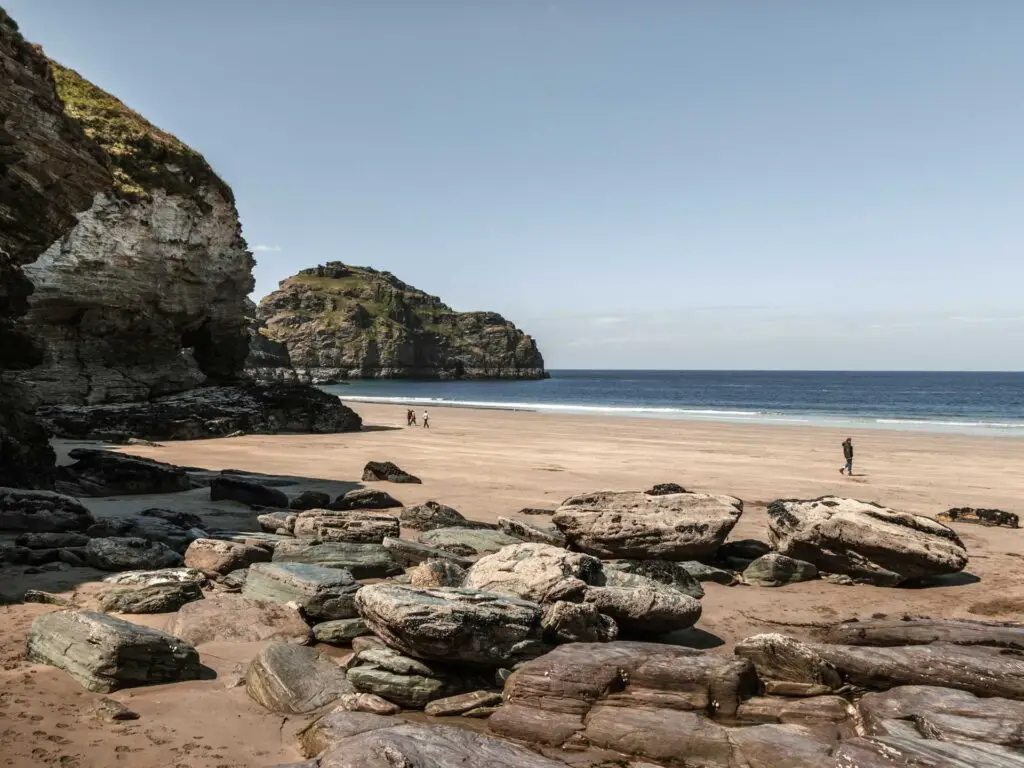 Large rocks leading to the sand beach of Bossiney Bay with a few people walking on it. There are cliffs to the left of the beach. The sea is blue.