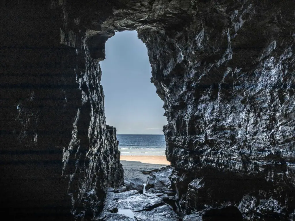 Standing inside a dark cave looking out to the beach and blue sea of Bossiney Bay. 