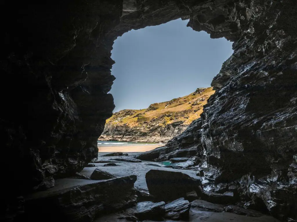 Standing in a cave on Bossiney Bay looking through the opening. The cave is dark and the view to the outside is of the sand, sea and cliffs.