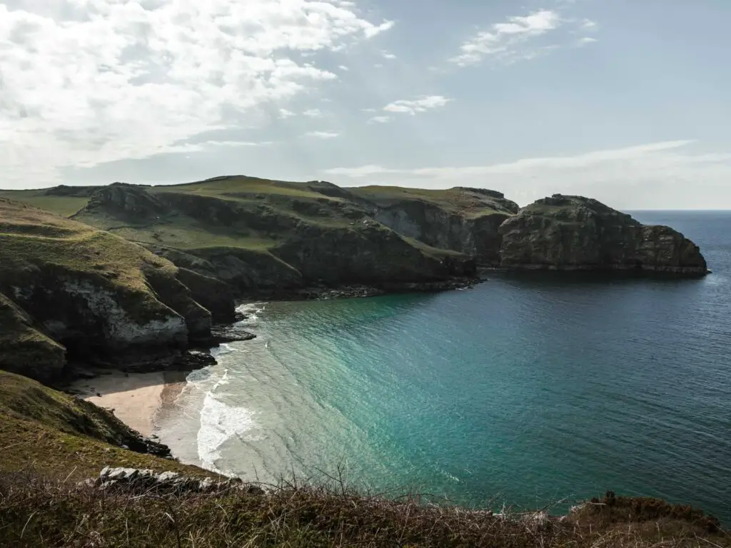 Looking down to the rugged cliff coastline and sand beach of Bossiney Bay on the walk from Tintagel. The sea goes from blue to turquoise as it meets the beach.