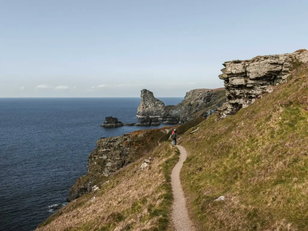 A narrow trail running along the side of a grass hill on the coastal Tintagel Bossiney circular walk. There are people at th end of the trail and rocks in the blue sea.