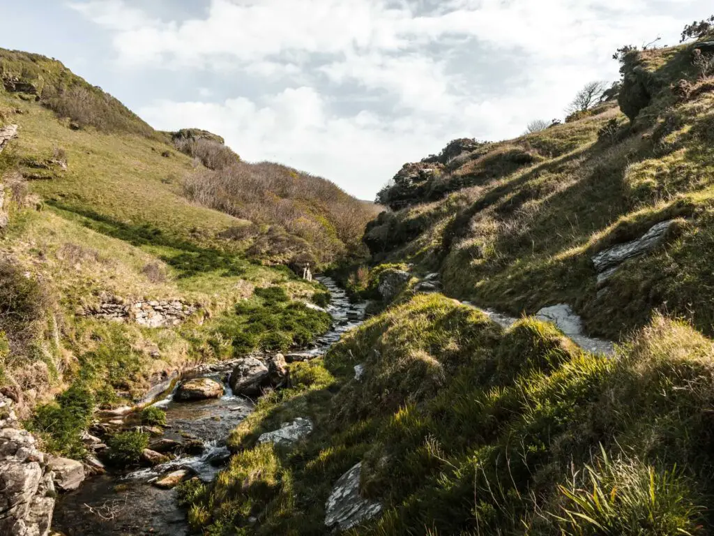 A stream running though the valley and a trail on the right side. The valley is enclosed by grass covered rugged hills.