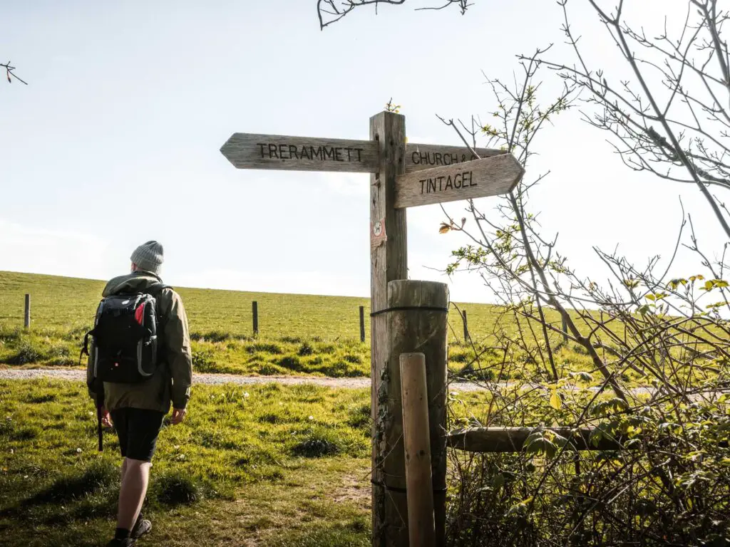 A man walking next to a wooden signpost pointing to Tintagel. There is a green field on the other side.