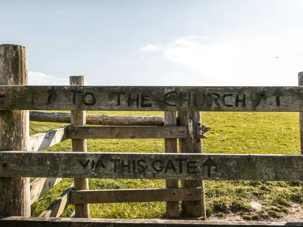 A wooden gate with writing pointing to the church. There is a green field on the other side.