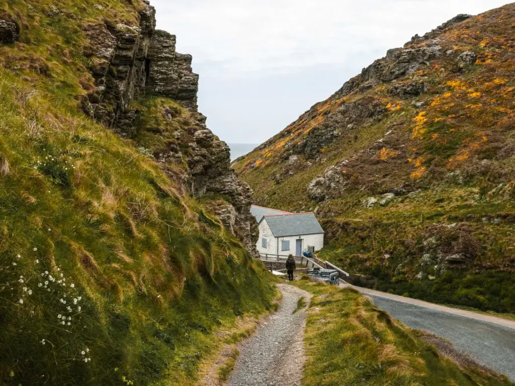 A gravel trail going downs the side of a hill on the coastal walk from Tintagel to Bossiney. There is a white building at the end of the trail between two hills. There is a man on the end of the trail.