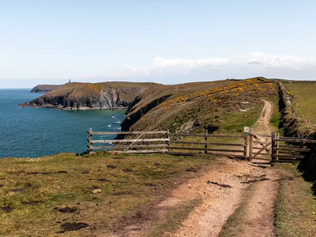 A dirt trail running along the grassy clifftop with Stepper Point in the distance, on the walk from Trevone and Gunver Head. There is a wooden fence across the trail.
