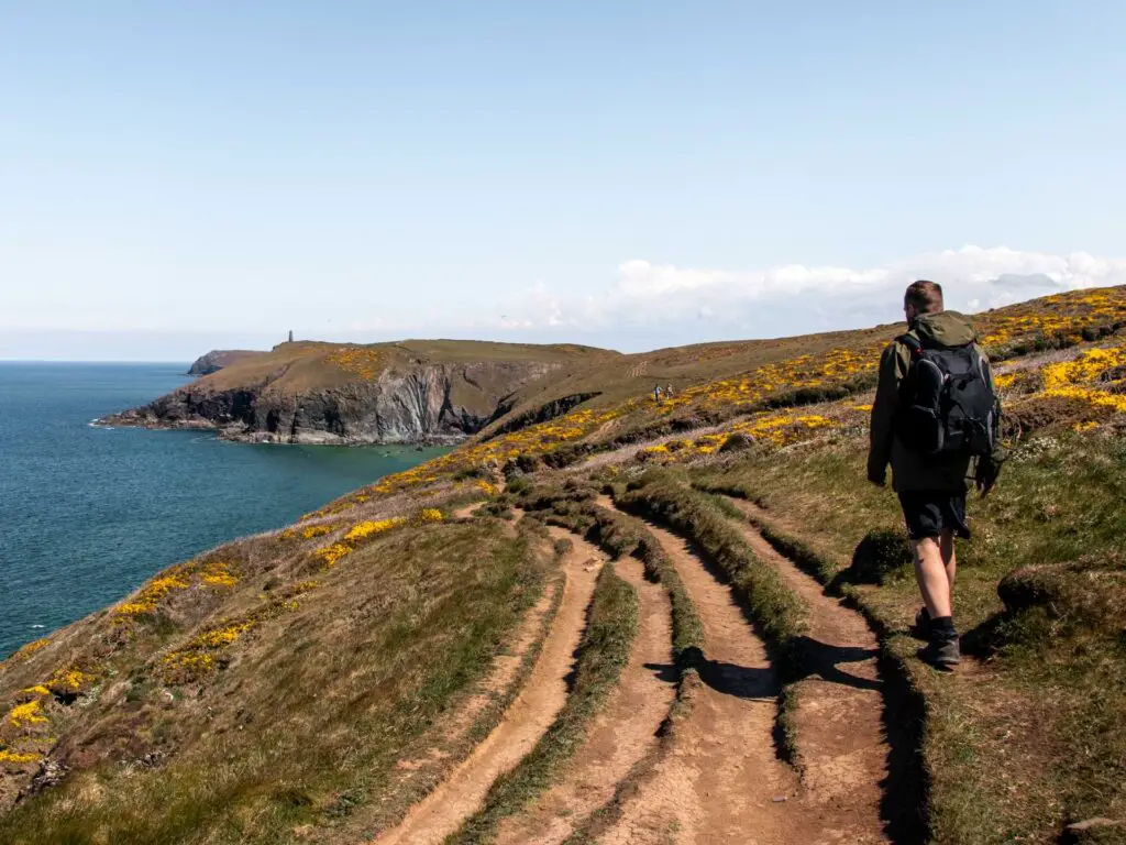 A dirt trail running through the grass clifftop with Stepper point in the distance on the walk from Trevone and Gunver Head. There is a man walking next to the trail.