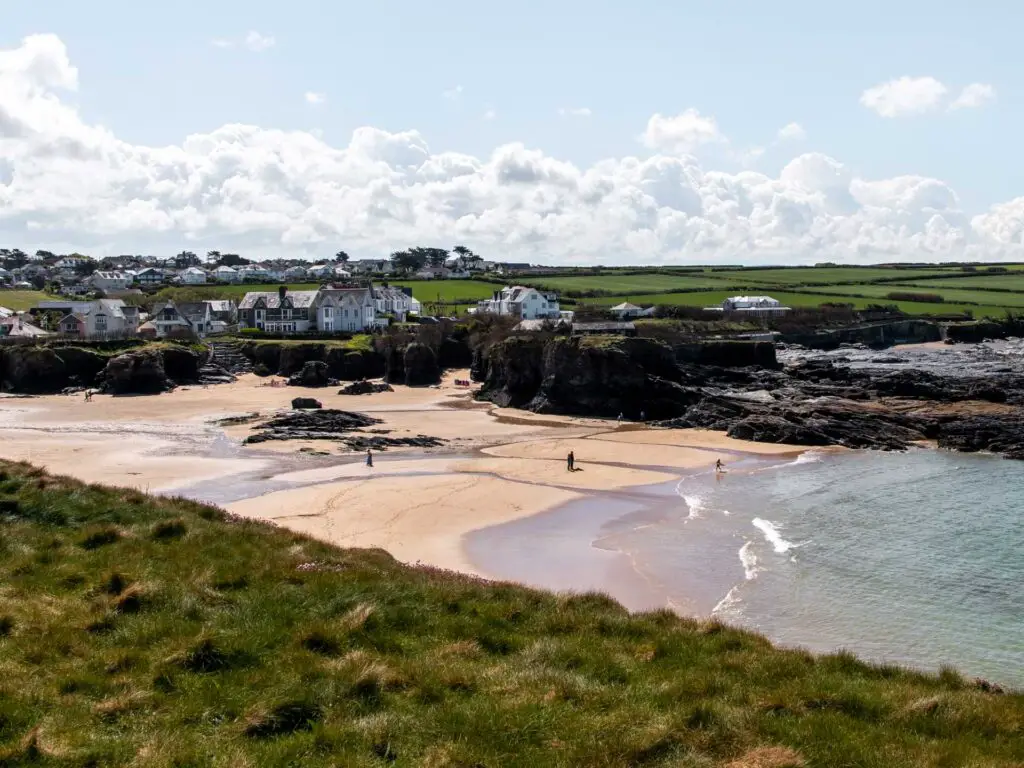 The pale coloured sandy beach of Trevone Bay. There are a few houses on the grass hill on the other side. There are a few people walking on the beach.
