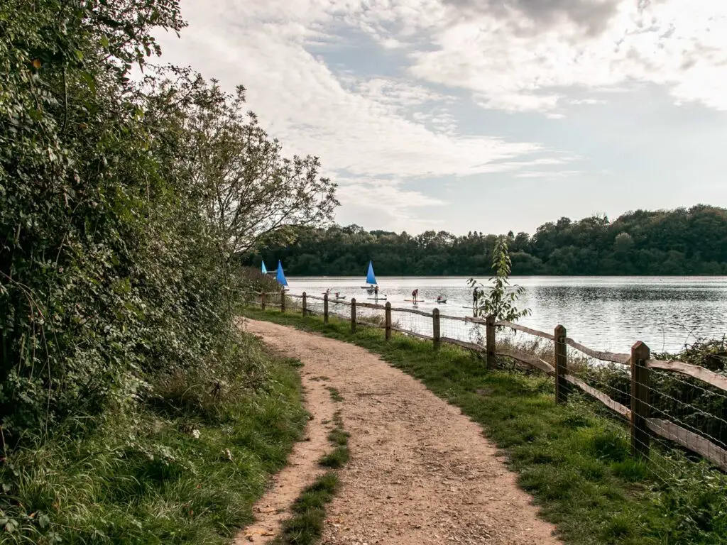 A gravel dirt trail to walk on, next to the Ardingly Reservoir as it curves around to the left. There are some sailboats and stand up paddle boarders on the water.