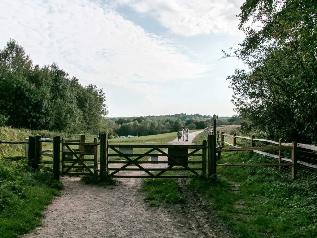 A gravel dirt trail leading to a wooden gate. There is a green field on the other side where the trail continues. There are a few people standing on the trail.