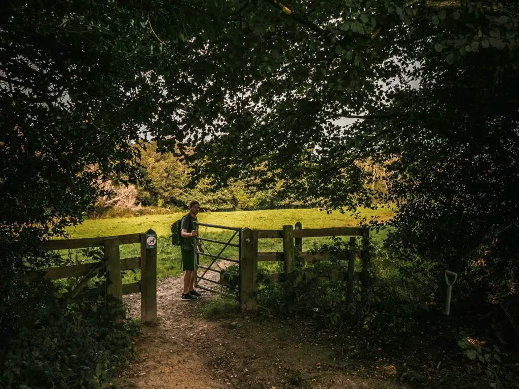 A man walking through a metal gate from tree cover to an open field on the circular walk from Balcombe to the ardingly reservoir and the ouse valley viaduct. The grass in the field is bright green with the light shining on it.