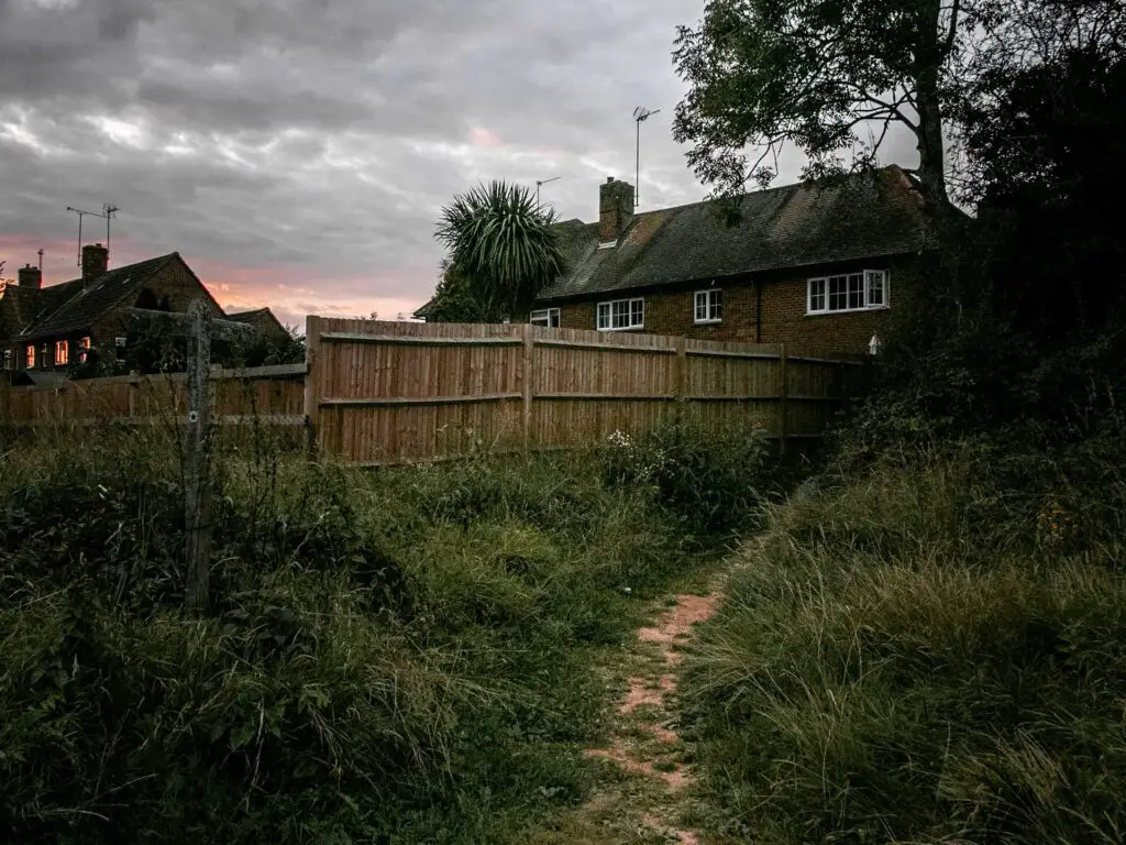 A small trail running alongside a fence , with a house partially visible on the other side of the fence. There is some pink in the sky from the setting sun.