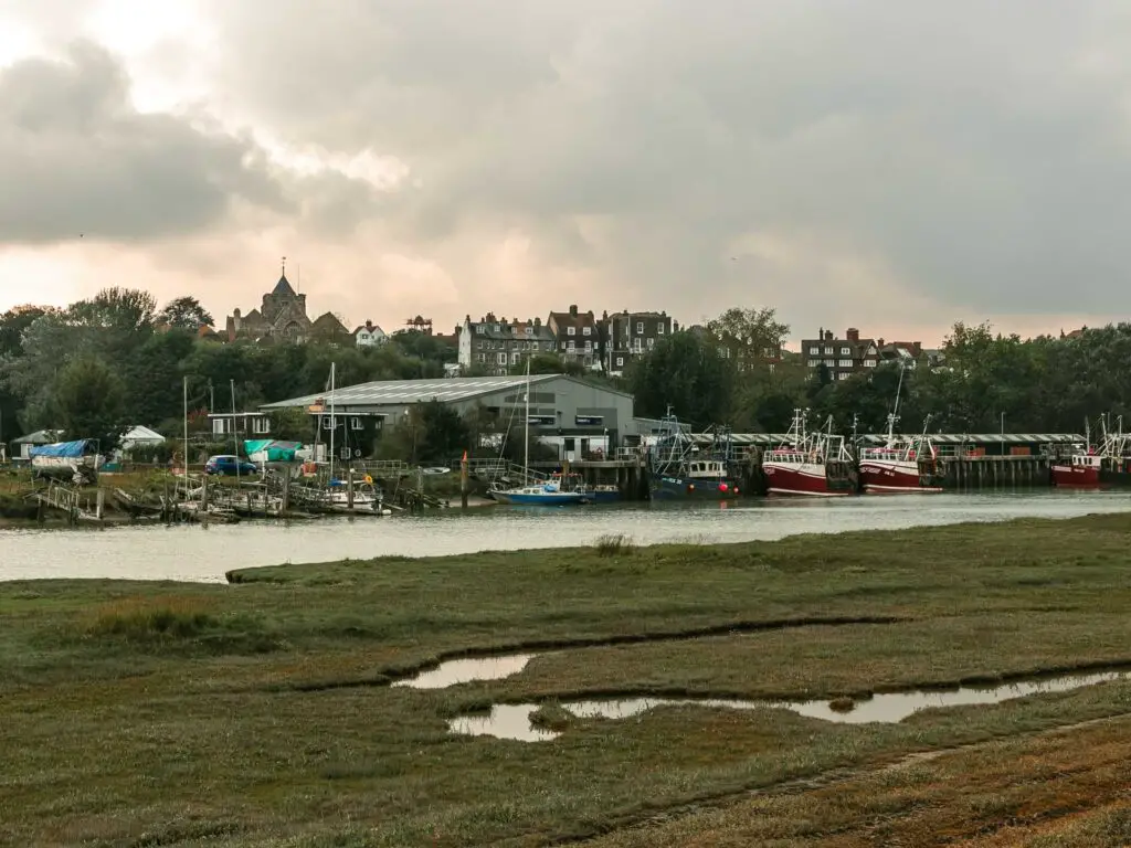 Marsh land with the River Rother on the other side. There are some boats moored along the river and tops of buildings of Rye visible in the distance. 