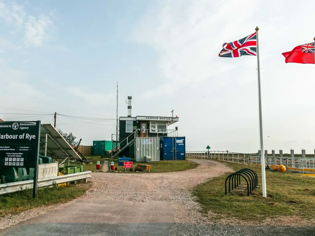 A gravel path leading to the Rye Harbour Master. There is a British flag raised on a flagpole.