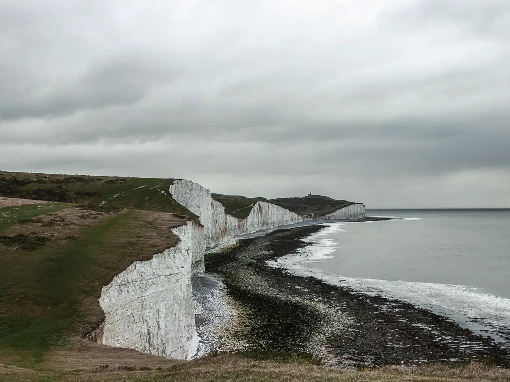 The white chalk cliffs looking back towards Birling Gap on the walk to Friston Forest. It is a grey gloomy day. The cliffs are toped with green grass.