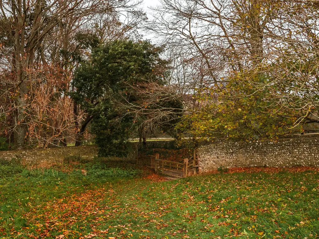 The side of the field enclosed by a brick wall and wooden gate. There are trees all along the wall.