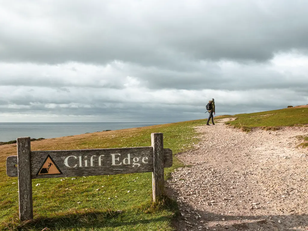 A wooden sign warning 'cliff edge' on the grass next to the gravel trail, on the circular walk away from Birling Gap. There is a man walking along the trail.