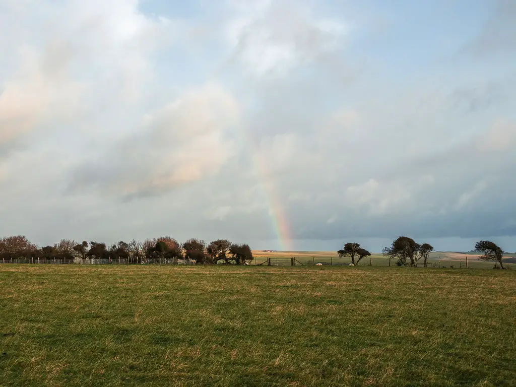 A rainbow coming up from the green field and across the cloudy blue sky on the circular walk back to Birling Gap.