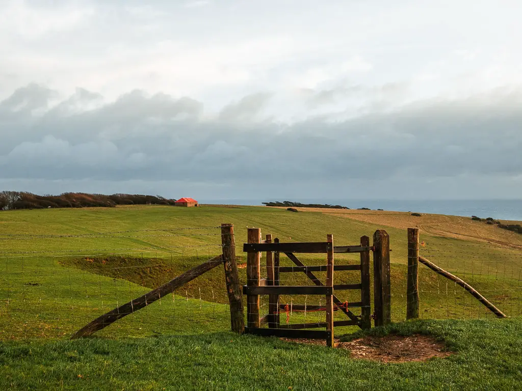 A wooden gate leading to an undulating green field, on the circular walk back to Birling Gap. There is a red roofed shed in the distance.