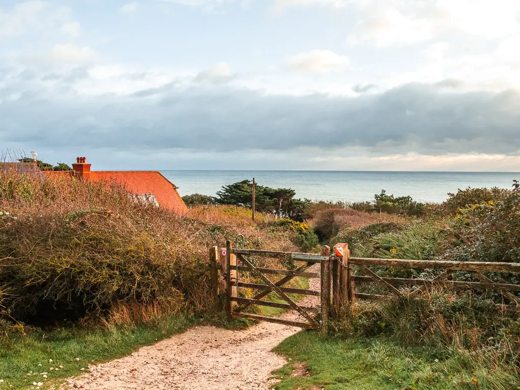 A wooden gate on a gravel dirt trail, leading through the bushes. A red roof of a house is peaking out from the bushes.