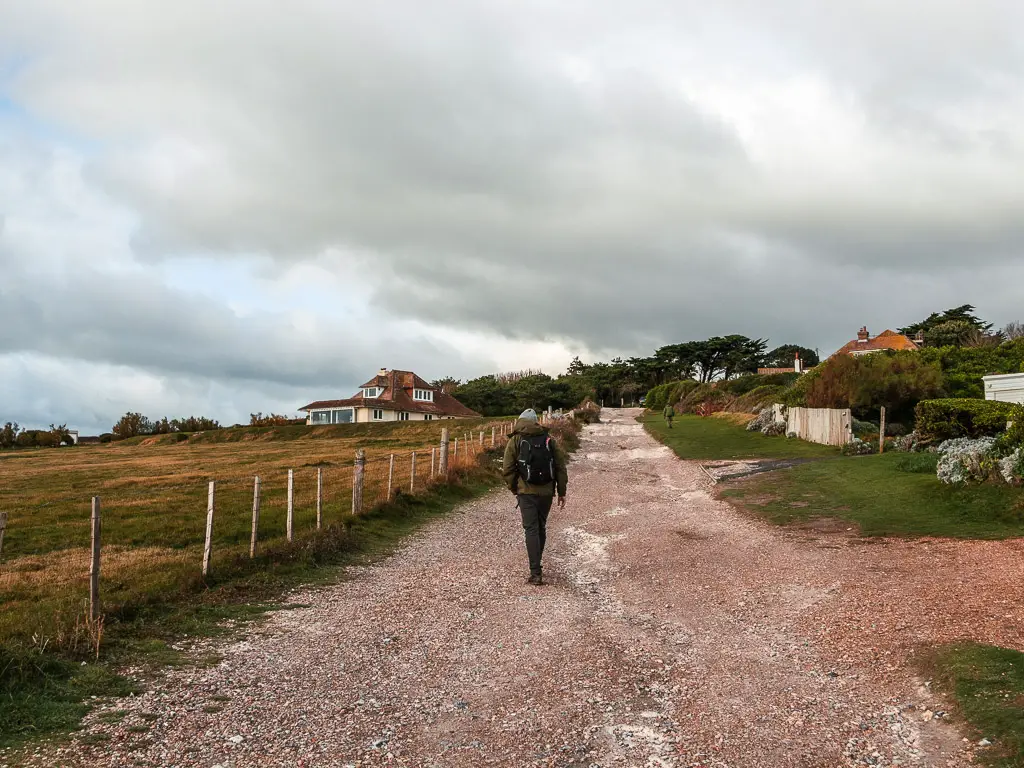 A man walking uphill along a gravel road on the walk out of Birling Gap. There is a green field on the left and small white house with red roof at the top. There are some houses on the right and trees ahead.