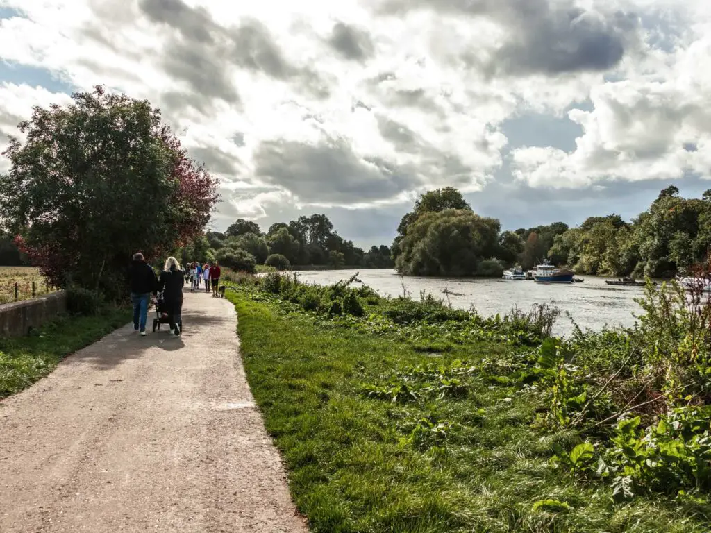 A wide path next to the River Thames. There is a green path of grass between the path and the river. There are a few people walking on the path.