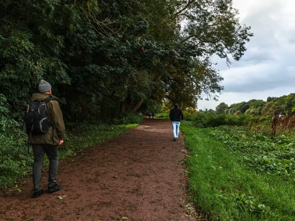 A dirt trail with two men walking on it. There is a green grass strip to the right and overhanging trees to the left.