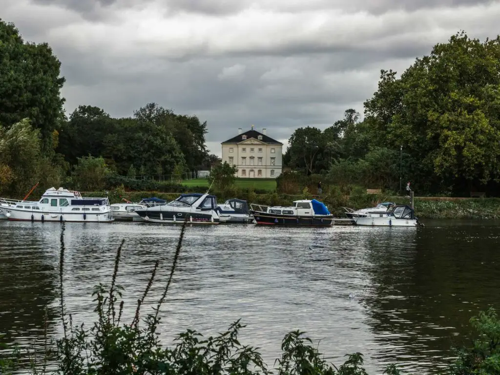 A group of boats moored on the side of the River Thames and a white building on the other side on the walk from Richmond to Hampton Court and bushy park.