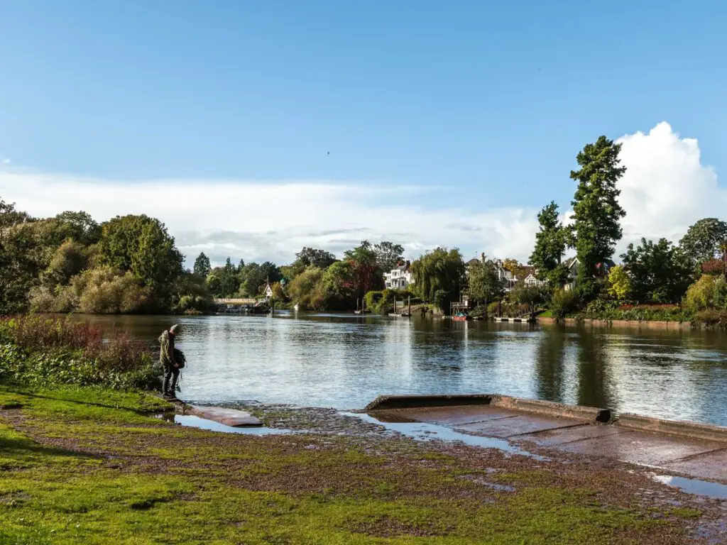 A man standing across the grass path next to the river. The other side of there river is lines with bushes and trees and a few houses poking through them.