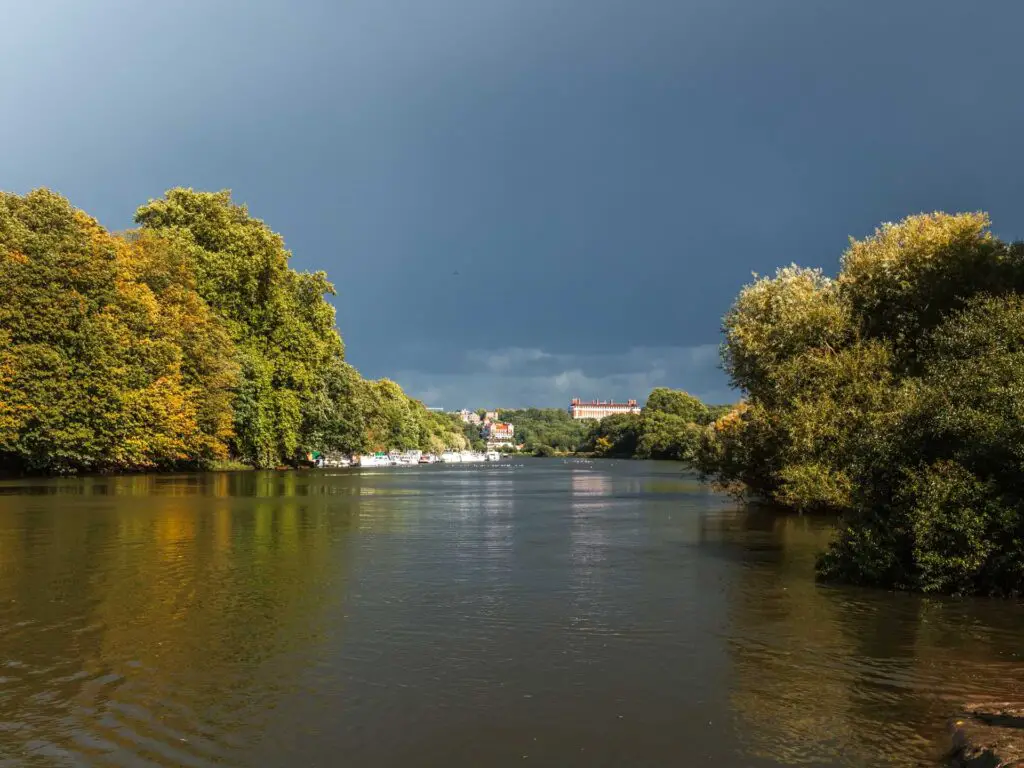 Looking down the River Thames towards Richmond, where the sky is dark grey on the walk towards Hampton Court. The river is lines with lots of green bushes and trees.