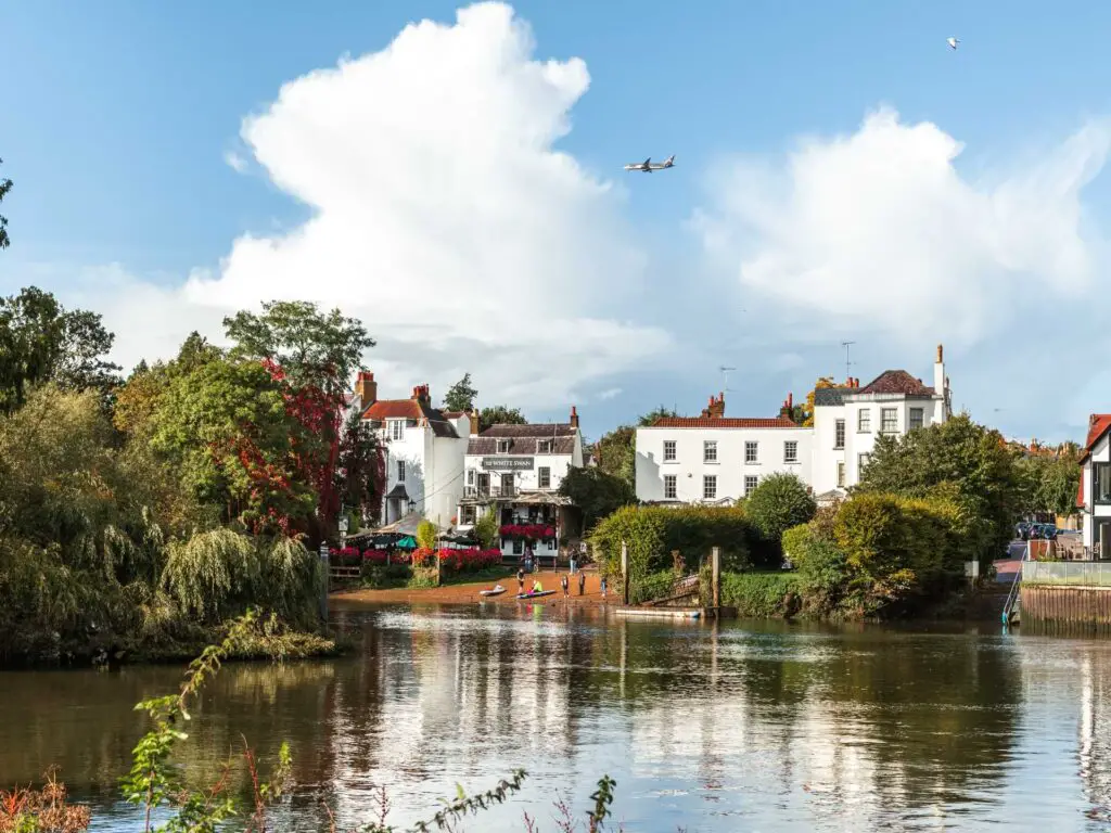 looking across the River Thames to an orange bank, lots of bushes and trees and some white buildings. The sky is blue with a few white clouds and a plane flying.