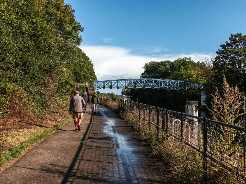 A path with bushes and trees to the left and a fence to the right. There is a blue metal bridge up ahead, and a few people walking on the trail.