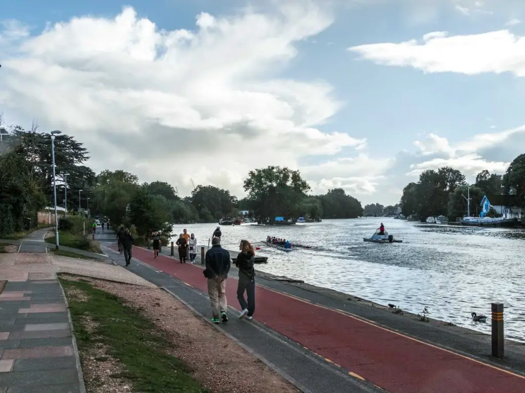 A red path next to the River Thames on the walk into Kingston. There are rowers on the river and a boat.