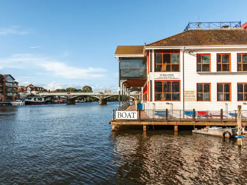 A white building with orange framed windows sitting on a platform on the river. There is a sign on the railing which says 'BOAT'. The railway bridge is visible past the building.