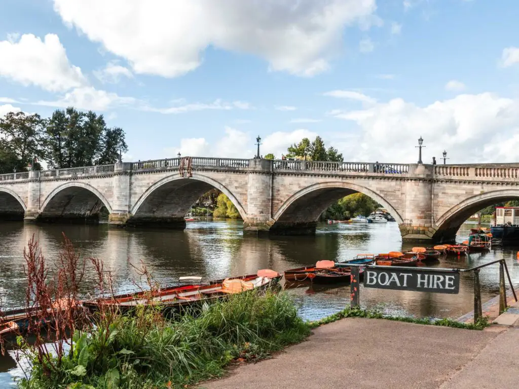 Looking onto the River Thames with Richmond bridge ahead. There is a sign saying 'boat hire' and a few row boats moored on the river.