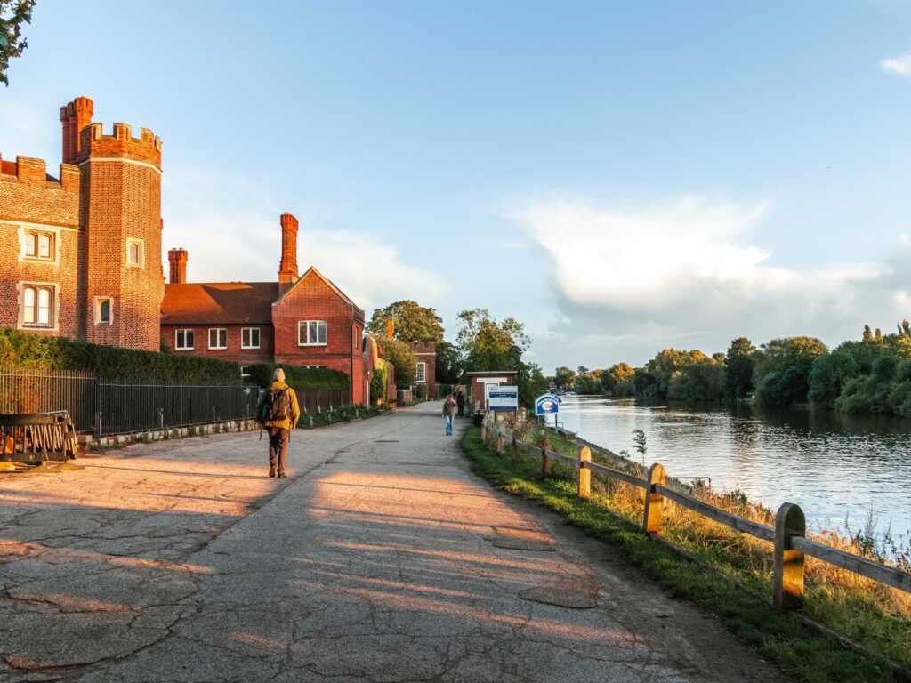 A wide path with buildings belonging to Hampton Court on the left, and the River Thames to the right. there is a man walking on the path.
