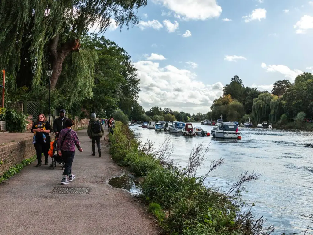 The Thames Path next to the river, with lots of people walking on it in Richmond. Ahead you can see lots of boats and trees lining the river.