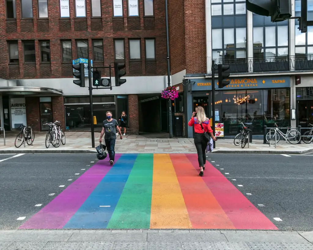 A rainbow coloured zebra crossing on the walk through Richmond. There are two people walking on the crossing. there are buildings and shops on the other side.