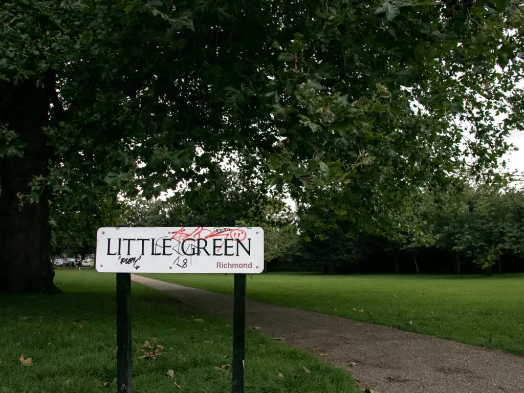 A white sign saying 'little green' with graffiti on it. The sign leads to a green grass field with a trail running through it.