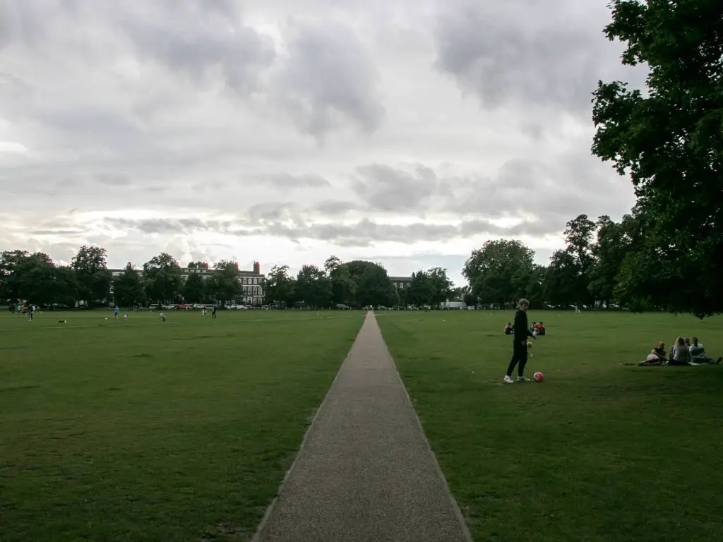 A long straight path through the green grass field. There are a fe people playing in the field.