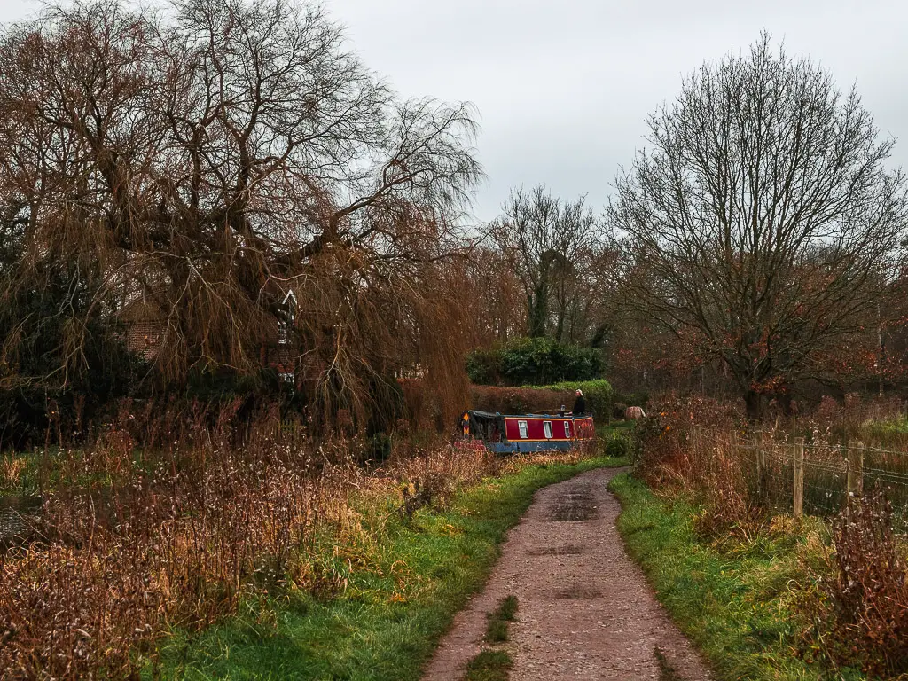 A trail lined with a grass bank next to the river. There is a red barge boat ahead, with a women standing on it. 