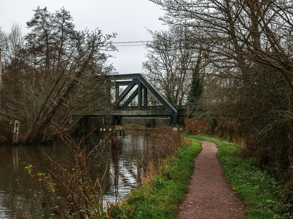 The dirt trail lines with grass next to the River Wey on the walk from Guildford to Godalming. There is a metal always bridge running over the river.